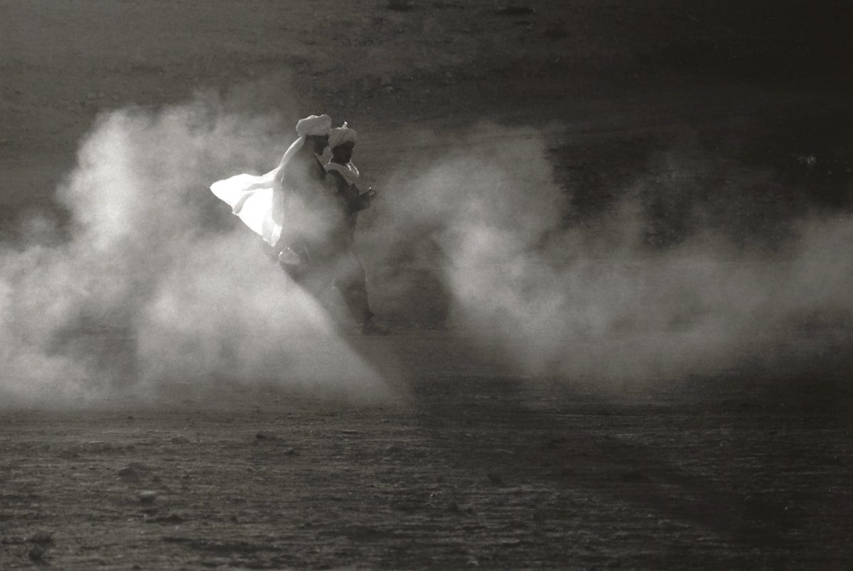 Black and white photograph of two people walkingthrough a dessert in a cloud of dust