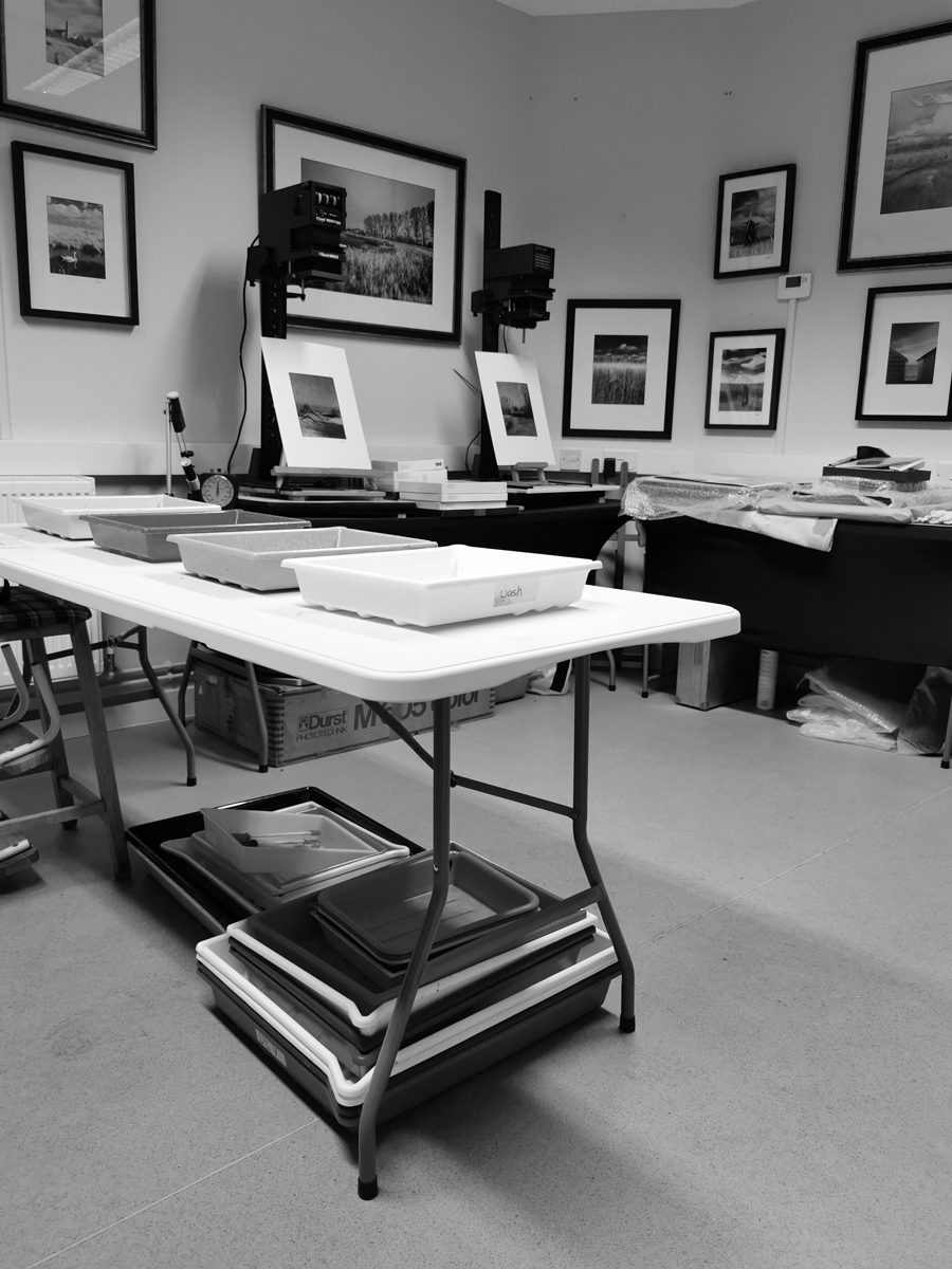 Black and white photograph of trays in the darkroom with photographs hung up on the wall in the background