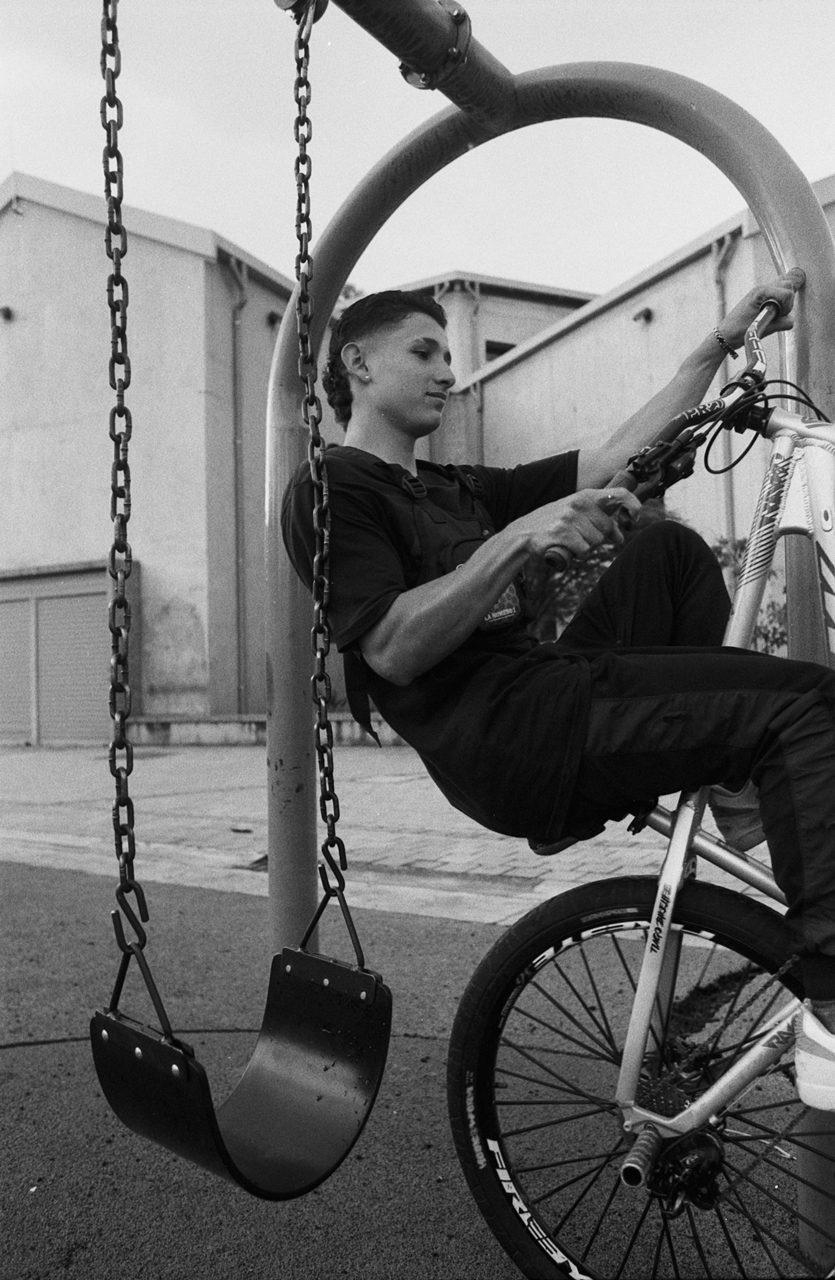 Henry Craver. Black and white shot of a guy doing a wheelie on a bike next to a swing.