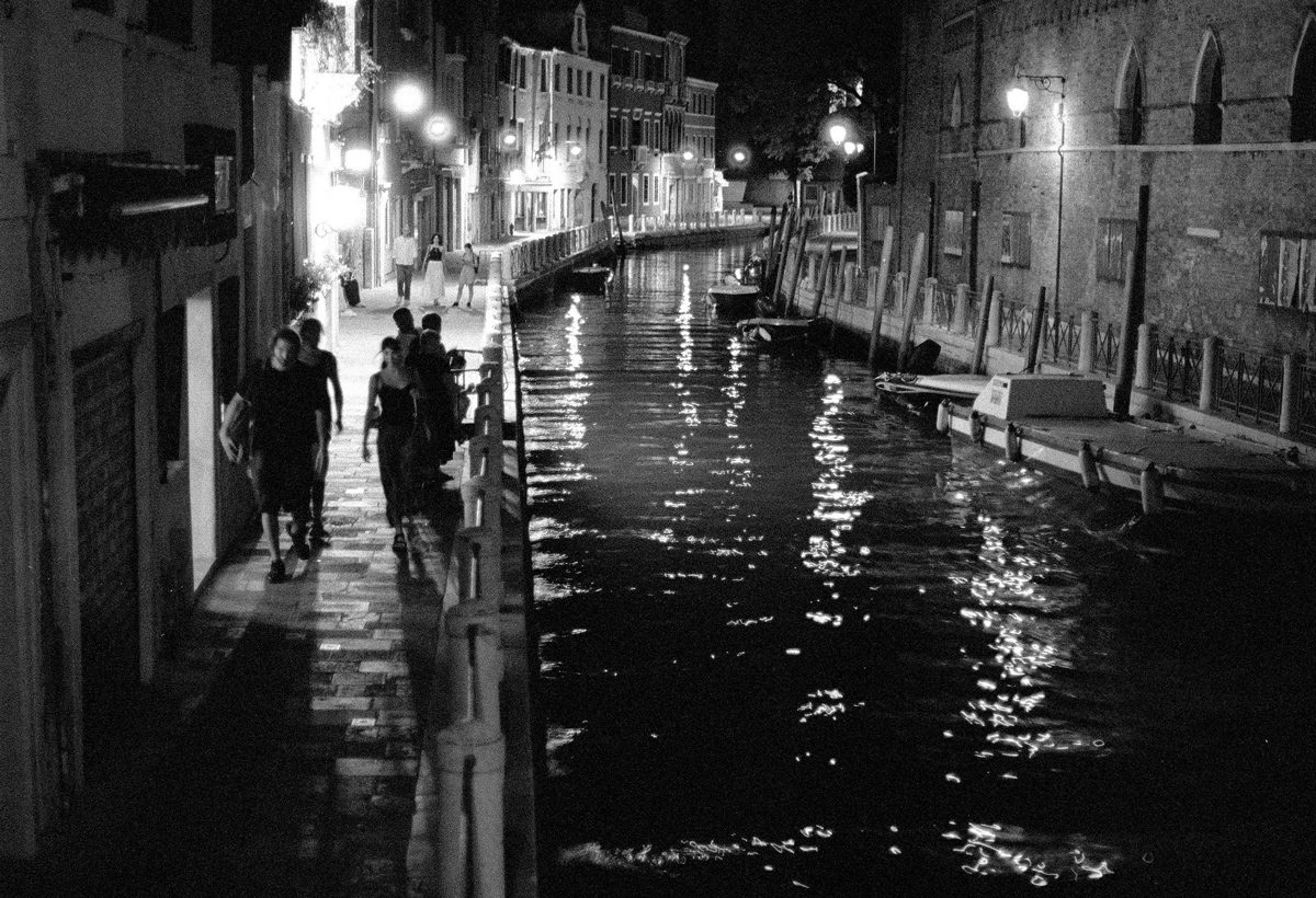Julian Tanase. Black and white night time shot in Venice of people walking along the canal.