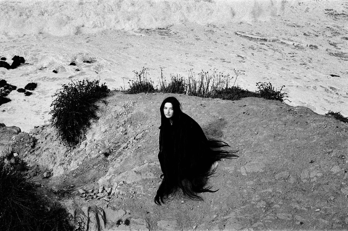 Black and white shot of a woman dressed in black on a rock in front of the sea
